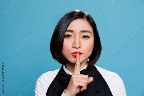 Hotel young asian woman receptionist making silent gesture, asking to keep secret portrait. Restaurant waitress wearing uniform holding forefinger on lips and looking at camera photo