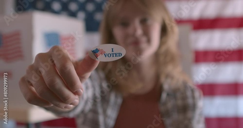 A white woman holding up an I voted sticker at a polling station with American Flag in the background  