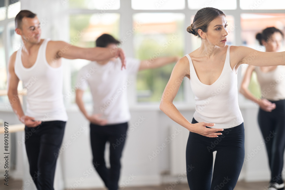 Experienced young female ballet dancer, dressed in white top and black leggings, executing dance moves with focused precision among group of people rehearsing in modern ballet studio