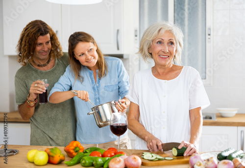 Cheerful couple and old woman drinking wine and talking with eath other while cooking in the kitchen at home photo