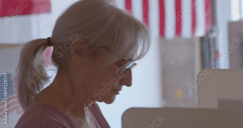 Close up of serious mature woman voting at a polling station with an American flag draped in the background