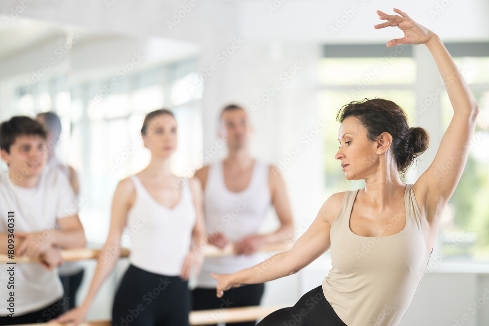 Professional female ballet instructor guiding focused group of amateur dancers standing at barre, demonstrating movement techniques in naturally lit dance studio..