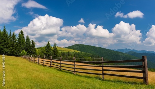 wooden fence on the meadow mountainous rural landscape of transcarpathia ukraine in summer carpathian countryside with forested rolling hill beneath a blue sky with white fluffy clouds