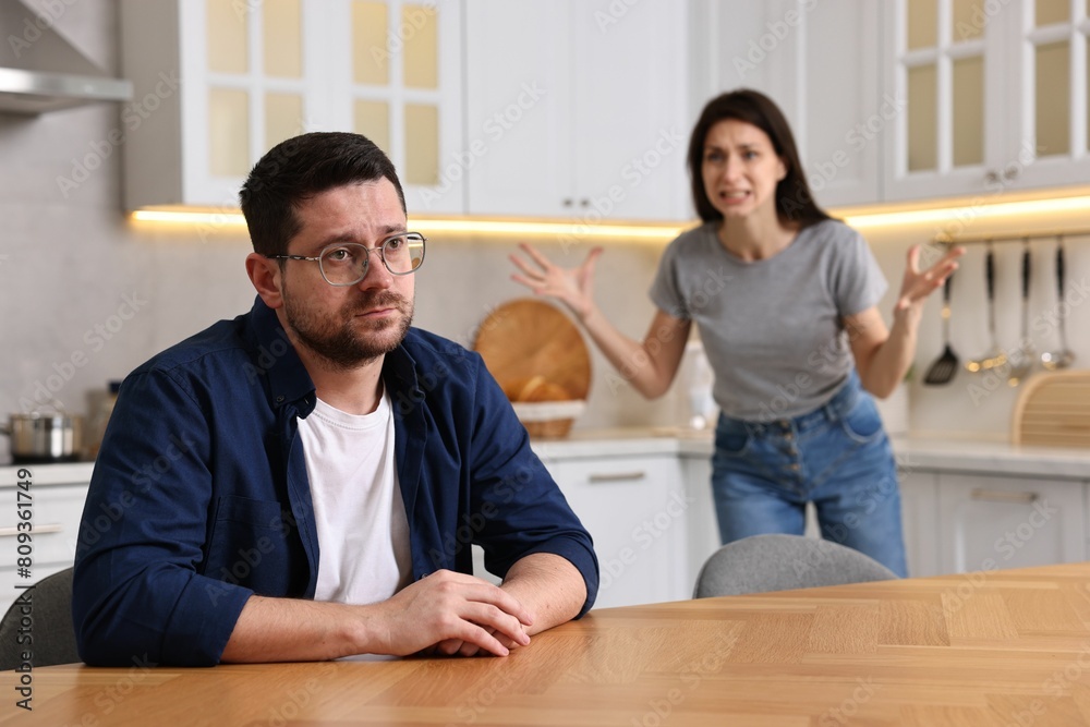 Obraz premium Sad husband sitting at table while his wife screaming at him in kitchen, selective focus. Relationship problems