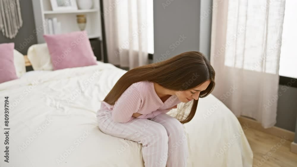 A pained young woman in pajamas clutches her stomach while sitting on a bed, indicating discomfort in a bedroom setting. A pained young woman in pajamas clutches her stomach while sitting on a bed, indicating discomfort in a bedroom setting.