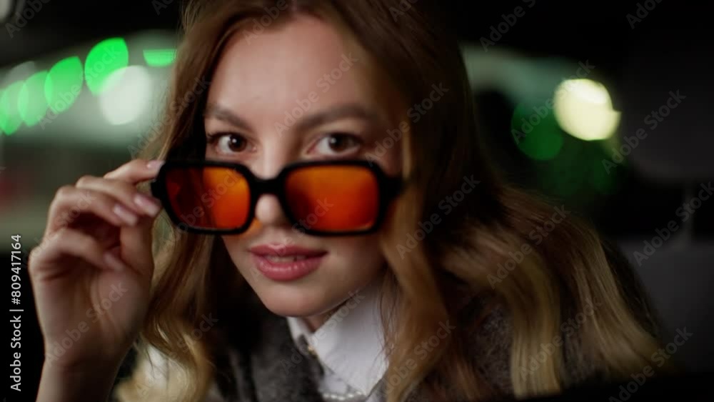 Woman in a car with orange-tinted sunglasses, gesturing with her hands as she talks. She is wearing a white shirt and a sweater, exuding a casual and expressive vibe inside a parked vehicle with green Woman in a car with orange-tinted sunglasses, gesturing with her hands as she talks. She is wearing a white shirt and a sweater, exuding a casual and expressive vibe inside a parked vehicle with green