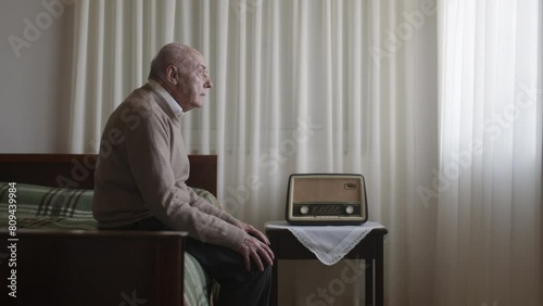 An elderly man sits on the edge of a bed in a softly lit room, a look of sadness on his face, with an old-fashioned radio beside him.