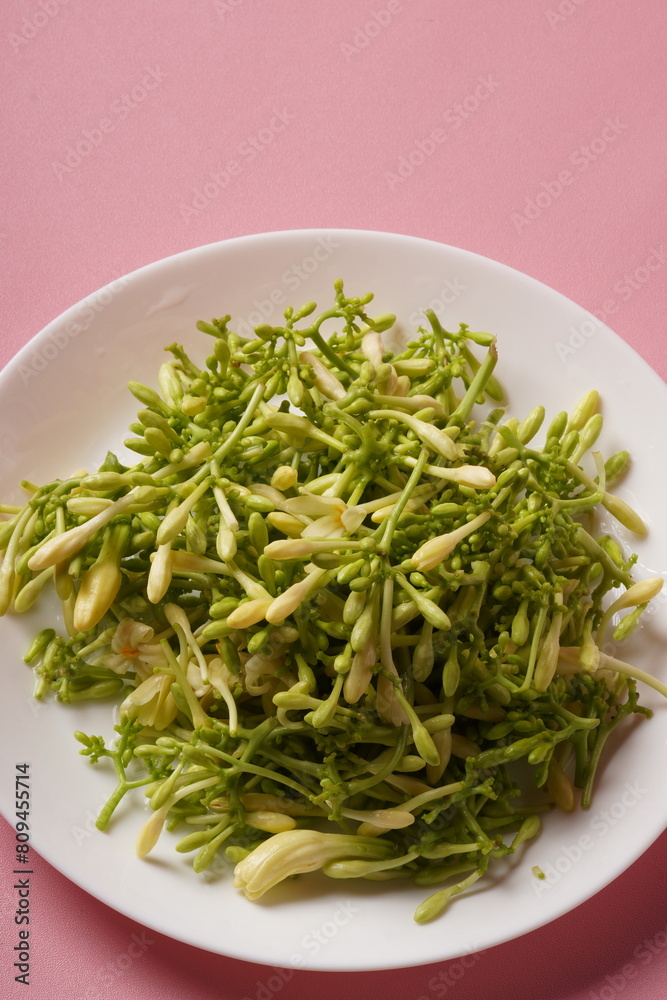 A pile of fresh, green, raw papaya flowers (Carica papaya), with mostly unopened buds, arranged on a white plate and photographed against a pink background. A pile of fresh, green, raw papaya flowers (Carica papaya), with mostly unopened buds, arranged on a white plate and photographed against a pink background.