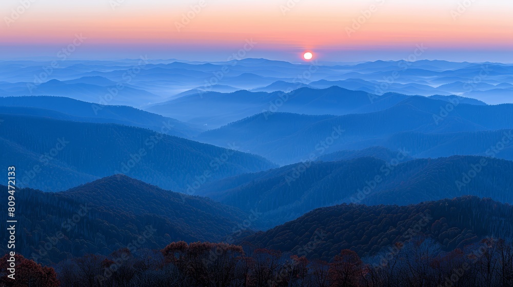   The sun sets over the distant mountains, with a valley and forested foreground in view