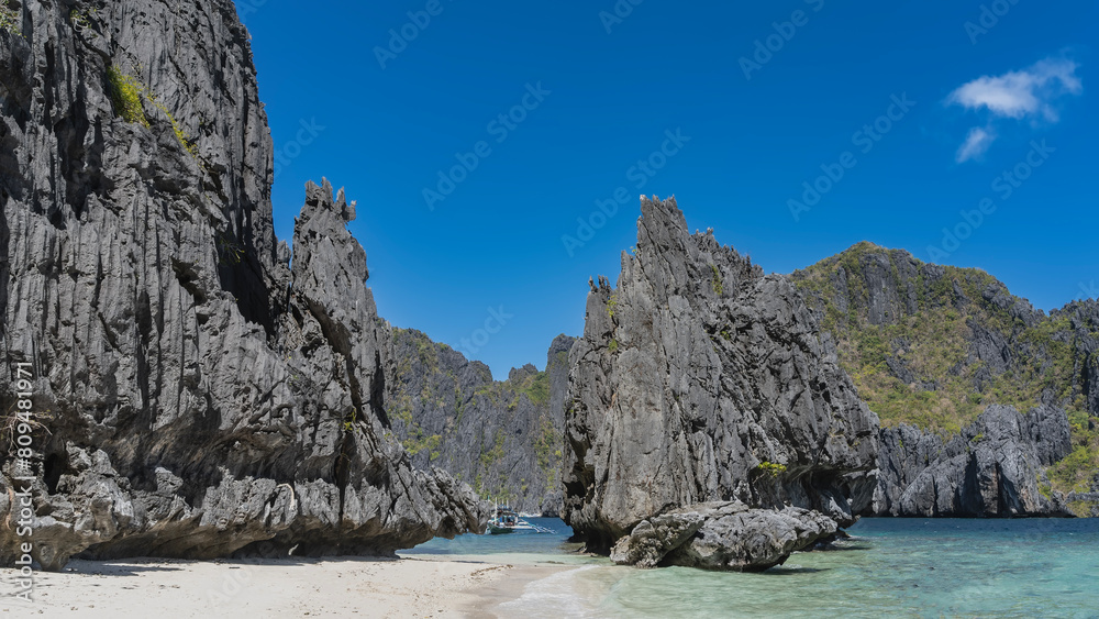 Naklejka premium Bizarre karst rocks with steep slopes surround the turquoise lagoon. A traditional Filipino bangka boat is visible between the cliffs. Sandy beach. Clear blue sky. Philippines. Palawan. Secret lagoon.