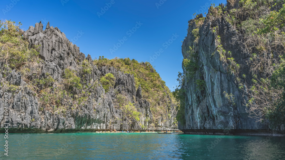 Many canoes with tourists sail along the emerald bay, surrounded by sheer karst rocks. Tropical vegetation on steep slopes. Clear blue sky. Philippines. Palawan. Bacuit Bay. The Big Lagoon. El Nido. Many canoes with tourists sail along the emerald bay, surrounded by sheer karst rocks. Tropical vegetation on steep slopes. Clear blue sky. Philippines. Palawan. Bacuit Bay. The Big Lagoon. El Nido.