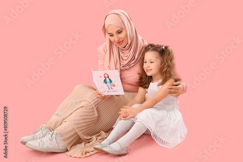 Little girl and her Muslim mother with greeting card sitting on pink background