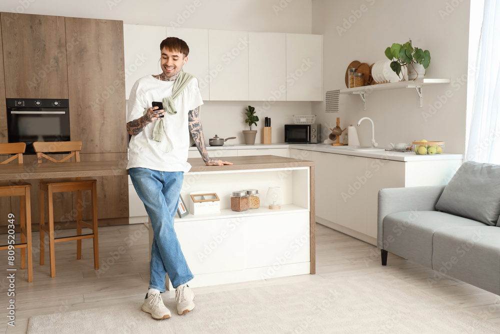 Young tattooed man with earphones and mobile phone listening to music in kitchen Young tattooed man with earphones and mobile phone listening to music in kitchen