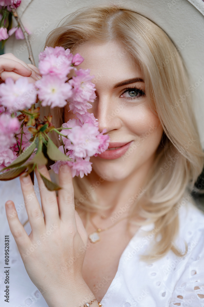 Beautiful young blonde woman in a hat holding a sakura branch in front of her
