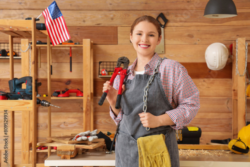 Young female carpenter with chain, bolt cutter and USA flag in workshop. Labor Day celebration