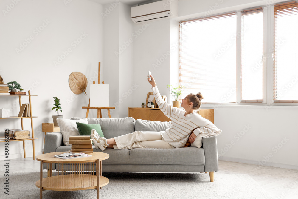 Young woman turning on conditioner with remote control and lying on sofa in living room Young woman turning on conditioner with remote control and lying on sofa in living room