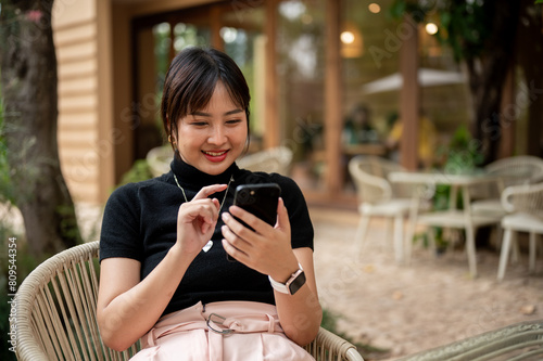 A cheerful, happy Asian woman sits at an outdoors table in the garden of a cafe using her smartphone