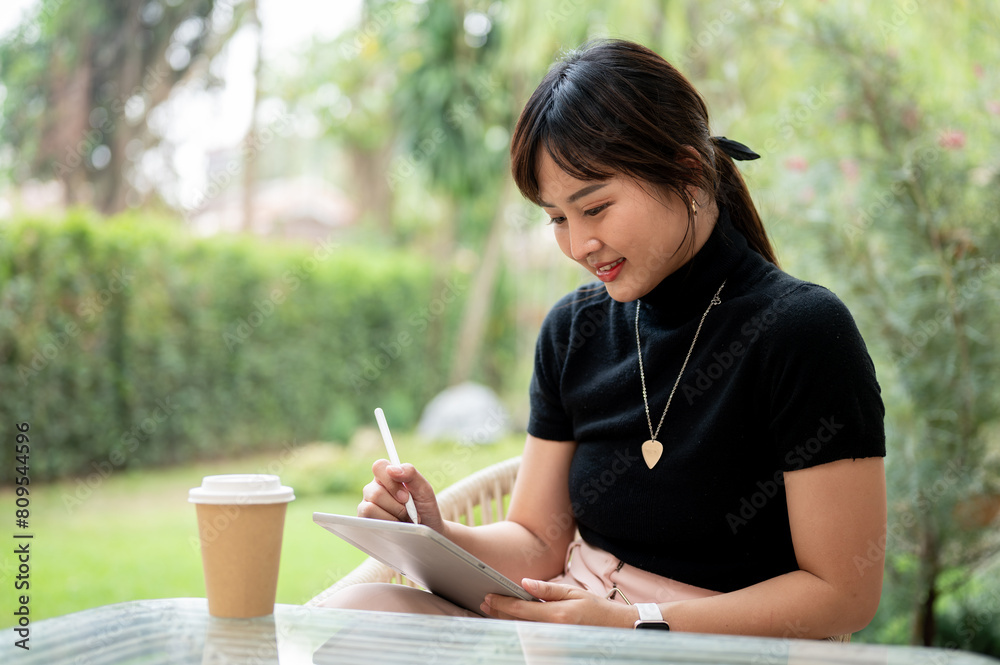 An attractive Asian woman sits at an outdoor table in a garden or backyard using her digital tablet. An attractive Asian woman sits at an outdoor table in a garden or backyard using her digital tablet.