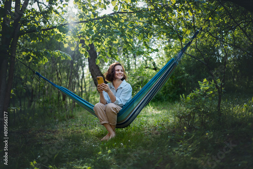 Cheerful young Caucasian woman in a hammock in a summer garden using a smartphone