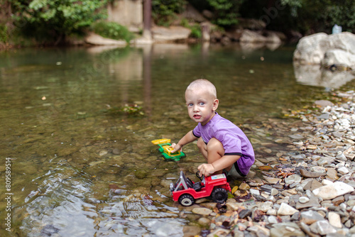 A little boy sits on the river bank, looks at the camera and holds toy cars. Dangerous games of children without parental supervision on the river bank. Child on the river