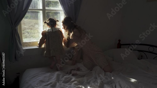A young girl looks out a window at a village sunrise, while her sister sits on the bed nearby, watching her carefully. photo