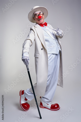 An elderly clown in a white coat, hat and large red patent leather clown boots. Walking stick in hand.
