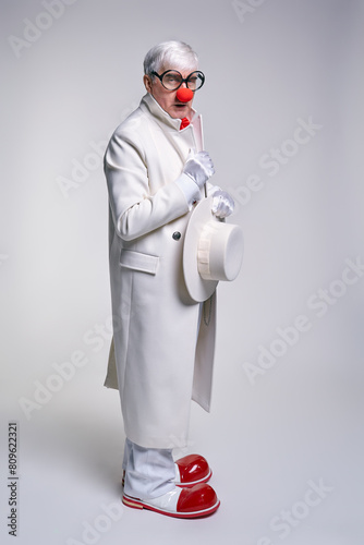 Serious, suspiciously looking clown in a white coat and large red patent leather clown boots with hat in hands.