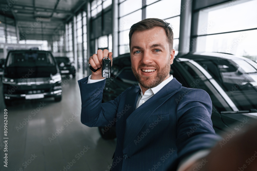 Making a selfie, with keys for new automobile. A businessman is in a car dealership Making a selfie, with keys for new automobile. A businessman is in a car dealership
