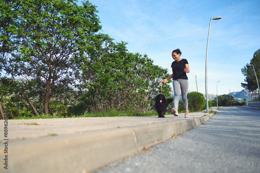 Front view of a young active woman walking her dog on leash in the nature on a beautiful sunny summer day. Pretty female athlete running with her pet in the morning