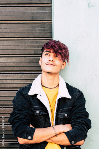 One young guy teenager standing against a wall and garage door looking on camera with trendy hair and denim jacket trucker looking up with confident expression. Modern teen male smiling