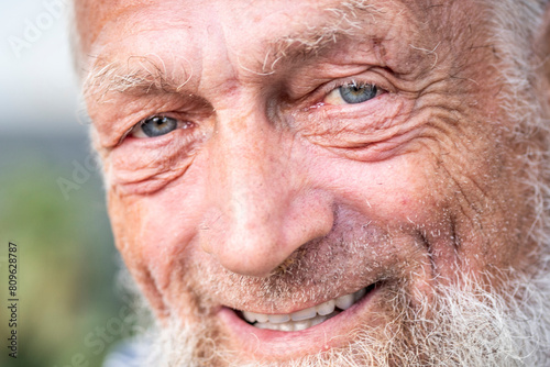 Close up portrait of happy 70-year-old optimist man with smiling wrinkled face, a touch of sadness feeling in his blue eyes. Park outdoors background defocused. Elderly people smile at the camera.