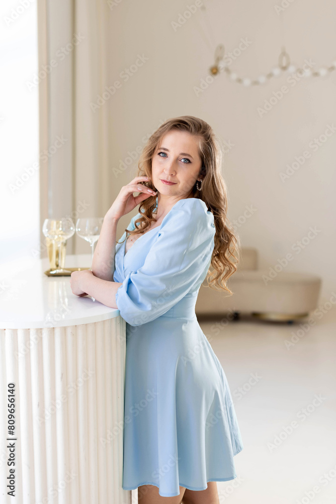 A young beautiful woman with fair hair wearing a blue dress is standing near a table with glasses... A young beautiful woman with fair hair wearing a blue dress is standing near a table with glasses...