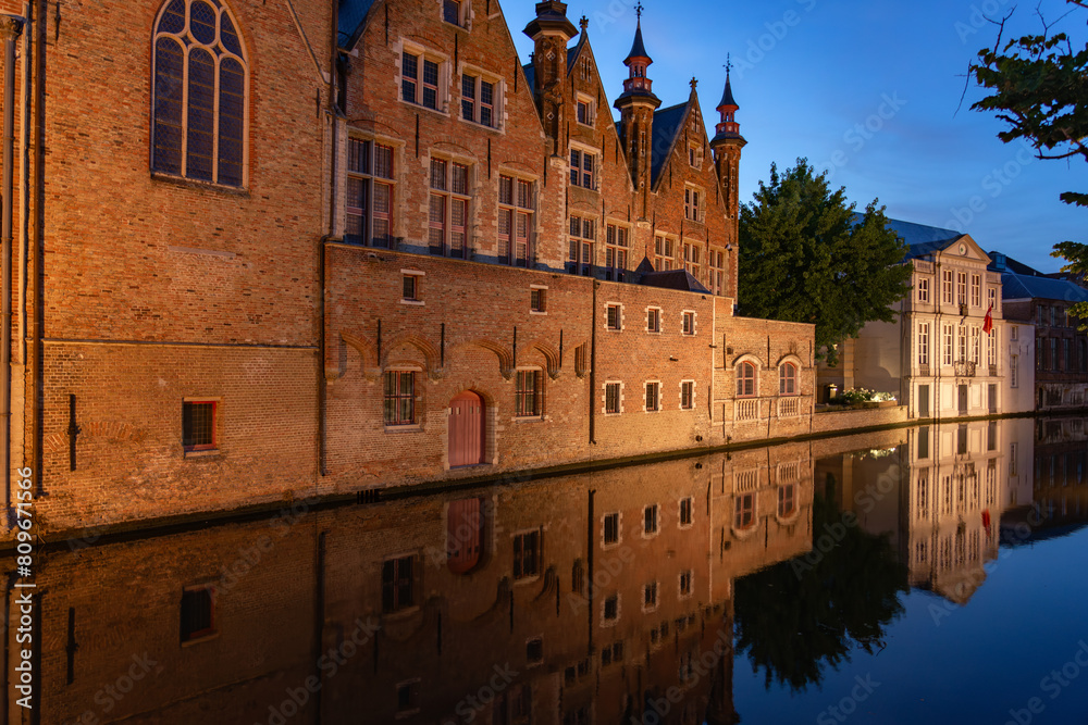 Historic buildings reflected on the canal in the old town of the beautiful city of Bruges in Belgium with the Meestrat bridge in the background at night.