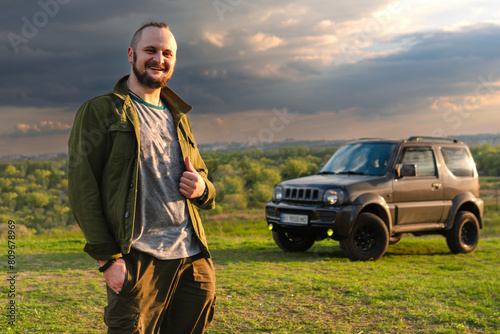 Happy traveler man near off-road vehicle in the background. The concept of active lifestyle and vacation road trip. A travelling by car to wilderness. Sunset clouds