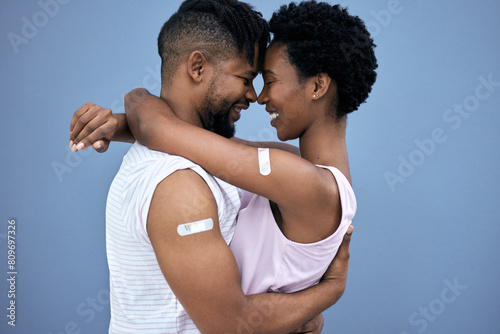 Vaccination, hug and black couple with plaster on arm in studio for cure, healthcare or booster shot on blue background. Smile, love and man with woman for medical injection, immunity or bandaid photo