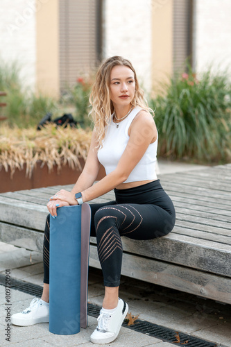 Positive young caucasian woman holding yoga mat after workout. Blonde girl wearing top and leggings sitting and looking away. Healthy lifestyle, keep fit concept