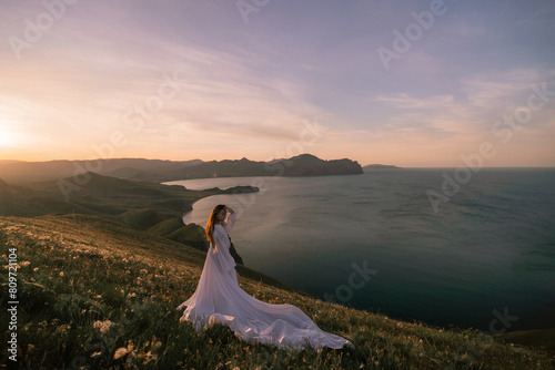 A woman stands on a hill overlooking the ocean in a long white dress © svetograph