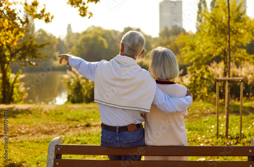 Old love is truest love. Romantic senior couple in love on date talking while sitting on park bench. Back view of mature man hugging his wife and showing her something from distance on warm evening. photo