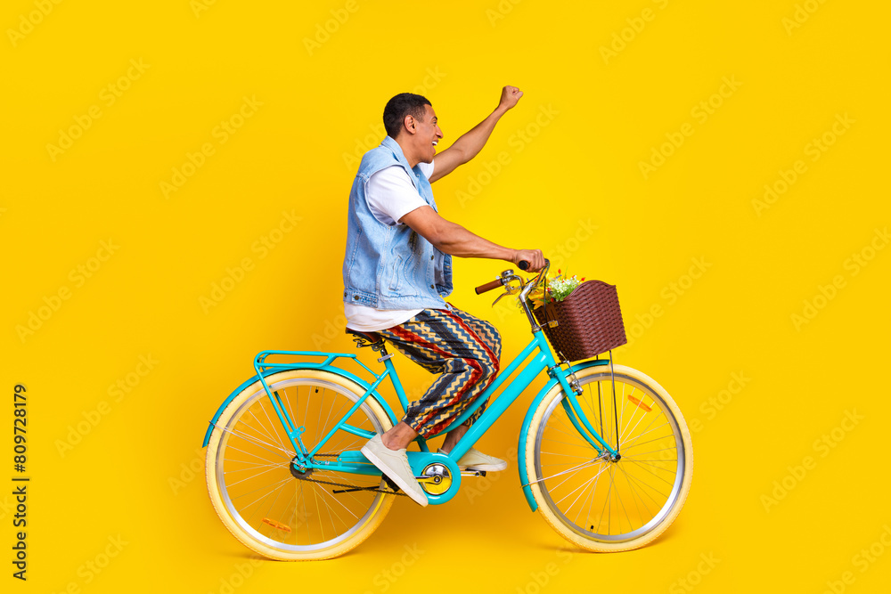 Full size photo of eccentric man dressed denim vest riding cycle to empty space raising fist up isolated on yellow color background Full size photo of eccentric man dressed denim vest riding cycle to empty space raising fist up isolated on yellow color background