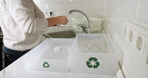 In the kitchen, a woman rinses plastic bottles and sorts them into a box with recycling signs. A woman removes caps from plastic bottles and sorts them into boxes for recycling. Eco-Friend. photo