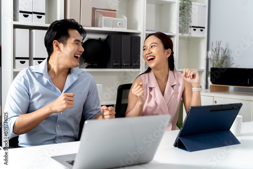Overjoyed Asian businessman and woman smiling and dancing with happiness and celebration of success while sitting on desk with computer laptop and tablet in the office. - Successful and fun workplace.