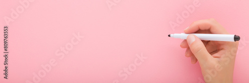 Young adult woman hand holding black white color pen and writing on light pink table background. Pastel color. Closeup. Wide banner. Empty place for text. Top down view. photo