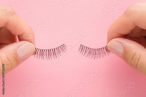Young adult woman hand fingers holding and showing new dark black false lashes on light pink table background. Pastel color. Female beauty product. Closeup. Point of view shot. Top down view.