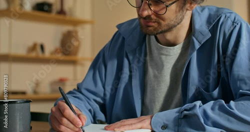 A man with glasses is writing in a notebook sitting at a table with a slight smile. Stylish home interior photo