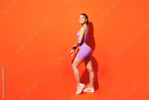 Full body side view young smiling fitness trainer instructor woman sportsman wear purple top clothes train in home gym look camera isolated on plain orange background. Workout sport fit abs concept.