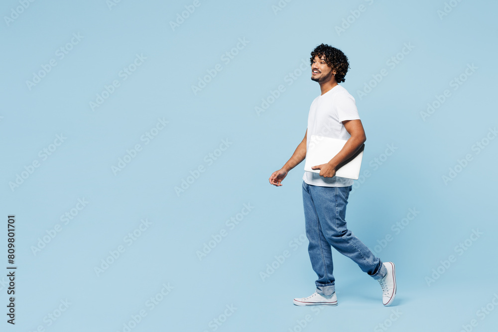 Full body side view young happy IT Indian man he wear white t-shirt casual clothes hold closed laptop pc computer walk go isolated on plain pastel light blue cyan background studio. Lifestyle concept.