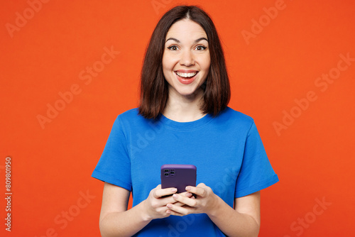Young smiling fun surprised happy woman she wear blue t-shirt casual clothes hold in hand use mobile cell phone look camera isolated on plain red orange background studio portrait. Lifestyle concept.