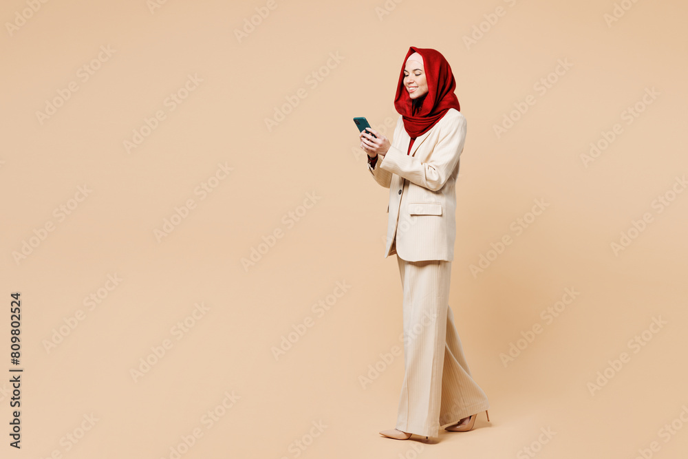 Full body young Arabian Asian Muslim woman she wear red abaya hijab suit clothes hold in hand use mobile cell phone walk go isolated on plain beige background studio. UAE middle eastern Islam concept.