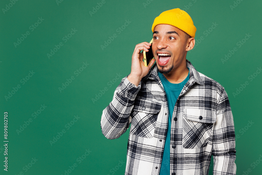 Young man of African American ethnicity he wears shirt blue t-shirt yellow hat talk speak on mobile cell phone conducting pleasant conversation isolated on plain green background. Lifestyle concept. Young man of African American ethnicity he wears shirt blue t-shirt yellow hat talk speak on mobile cell phone conducting pleasant conversation isolated on plain green background. Lifestyle concept.