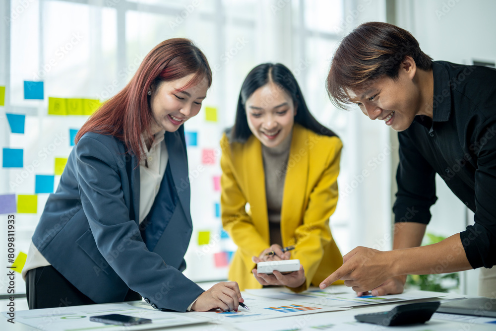 Three people are smiling and working together at a table. They are wearing business attire and have a whiteboard with colorful sticky notes on it Three people are smiling and working together at a table. They are wearing business attire and have a whiteboard with colorful sticky notes on it